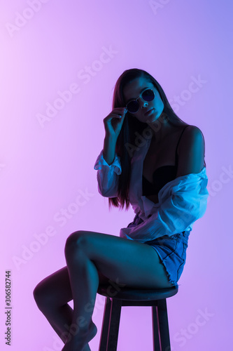 Young sexy woman in short and shirt, sunglasses sitting on the chair isolated on light background