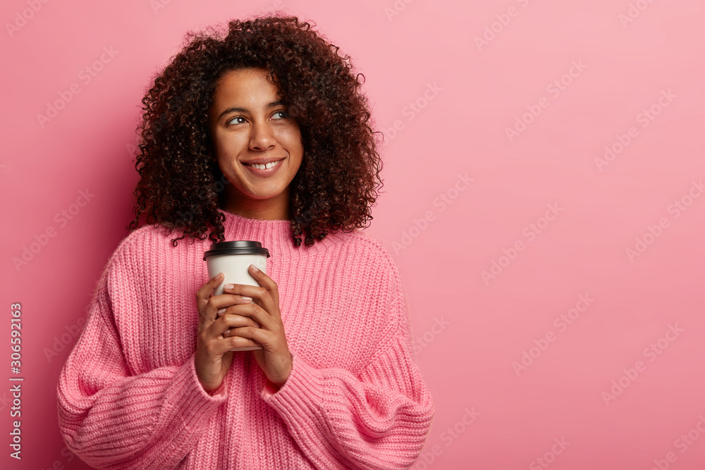Dreamy beautiful Afro American lady enjoys caffeine beverage, holds disposable cup with coffee, looks happily aside, wears pink sweater, enjoys rest, poses indoor. Student has break after classes Dreamy beautiful Afro American lady enjoys caffeine beverage, holds disposable cup with coffee, looks happily aside, wears pink sweater, enjoys rest, poses indoor. Student has break after classes