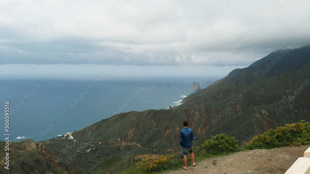 Aerial view. A single man stands on the edge of a mountain, looking at a beautiful view - the long coast of the Atlantic Ocean, the green valley and the mountains of the Anaga National Park, Tenerife