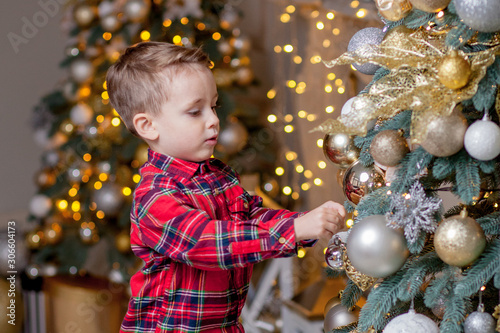 Happy little boy in elegant festive suit decorates beautiful Christmas tree at home. Merry Christmas and Happy New Year