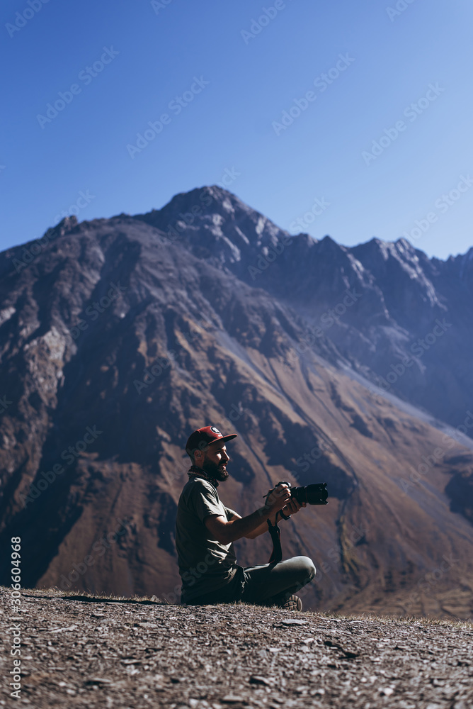 Fototapeta premium stylish bearded man with camera in hands on top of Georgian mountains: nature photographer traveler taking photo of beautiful landscape from top of the mountain