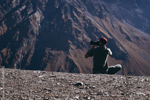 stylish bearded man with camera in hands on top of Georgian mountains  nature photographer traveler taking photo of beautiful landscape from top of the mountain