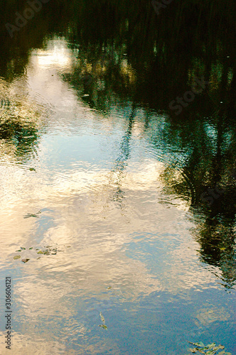Painting-like image of shadowy reflection of dark trees, light blue sky and scattered clouds and sunlight in the ripples of a pond just before sunset