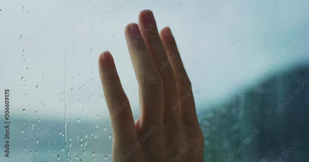 Close up of a young woman hand is touching a glass with water drops while outside is raining.