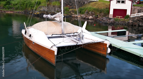 Wood hull catamaran sailboat tied to a dock. Rocky shore and mountain and buildings in background.