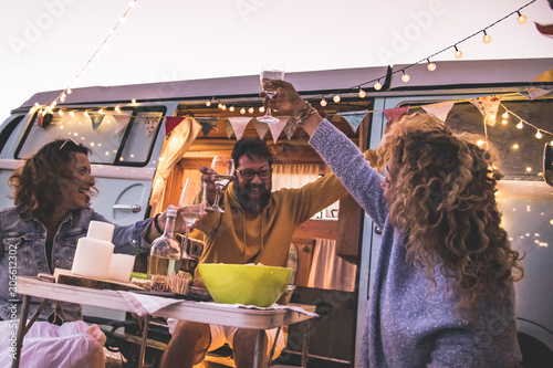 Happy friends cheering and drinking wine during camping vacation at the beach with vintage van. People having fun at weekend summer evening with camper. Youth lifestyle togetherness and party concept