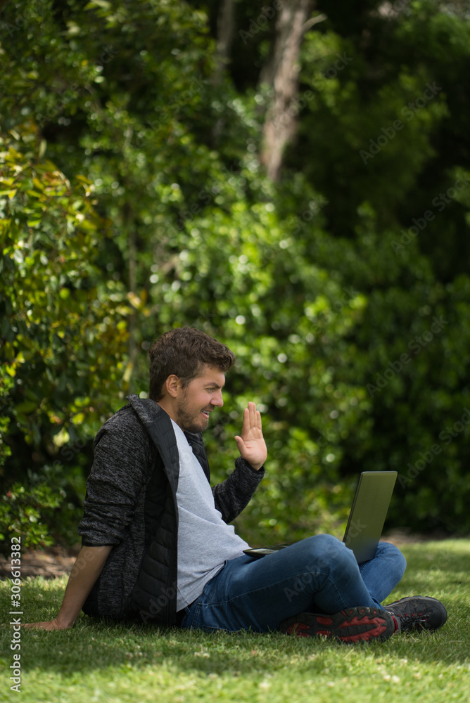 Man having a video call with his computer saying hello through the screen. He is smiling. Natural Environment. Vertical photography Man having a video call with his computer saying hello through the screen. He is smiling. Natural Environment. Vertical photography