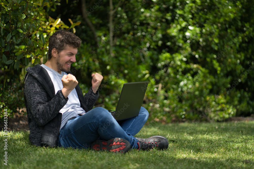 Man sitting on a park while watching his computer and seems to celebrate furiously with his hands. Natural Environment. Technological concept Man sitting on a park while watching his computer and seems to celebrate furiously with his hands. Natural Environment. Technological concept