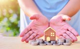 Concepts of protecting and helping the family and family debt problems. (There are hands protecting the pile of coins on the white wooden floor and the house model is placed together.)