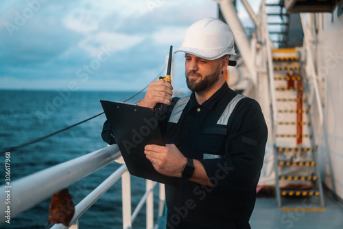 Marine Deck Officer or Chief mate on deck of offshore vessel or ship , wearing PPE personal protective equipment - helmet, coverall. He holds VHF walkie-talkie radio in hands. photo