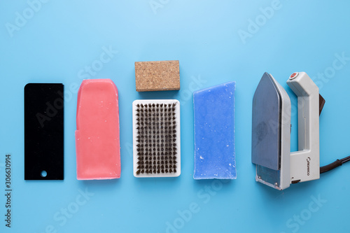 Equipment needed to wax skis including wax, iron, scraper, brush and cork styled on a plain blue winter background. Flat lay.