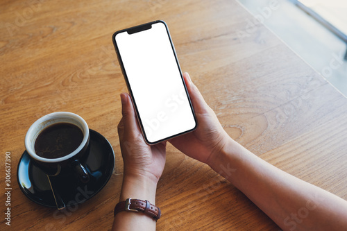 Mockup image of a woman holding black mobile phone with blank screen with coffee cup on wooden table