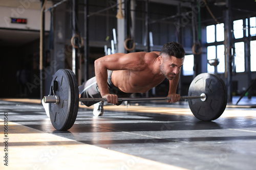 Fit and muscular man doing horizontal push-ups with barbell in gym.