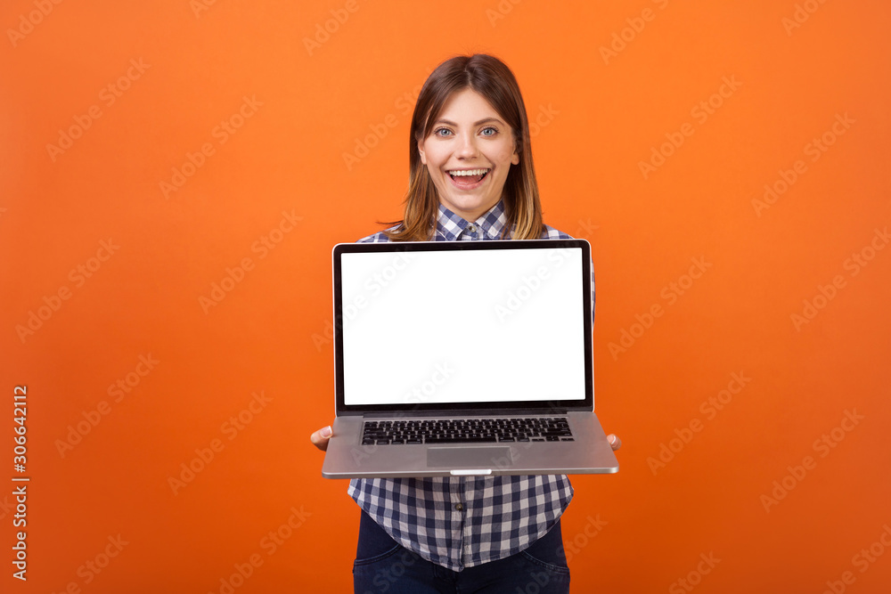 Naklejka premium Portrait of glad beautiful woman with brown hair in checkered shirt standing holding laptop with blank screen and smiling at camera, place for ad. indoor studio shot isolated on orange background