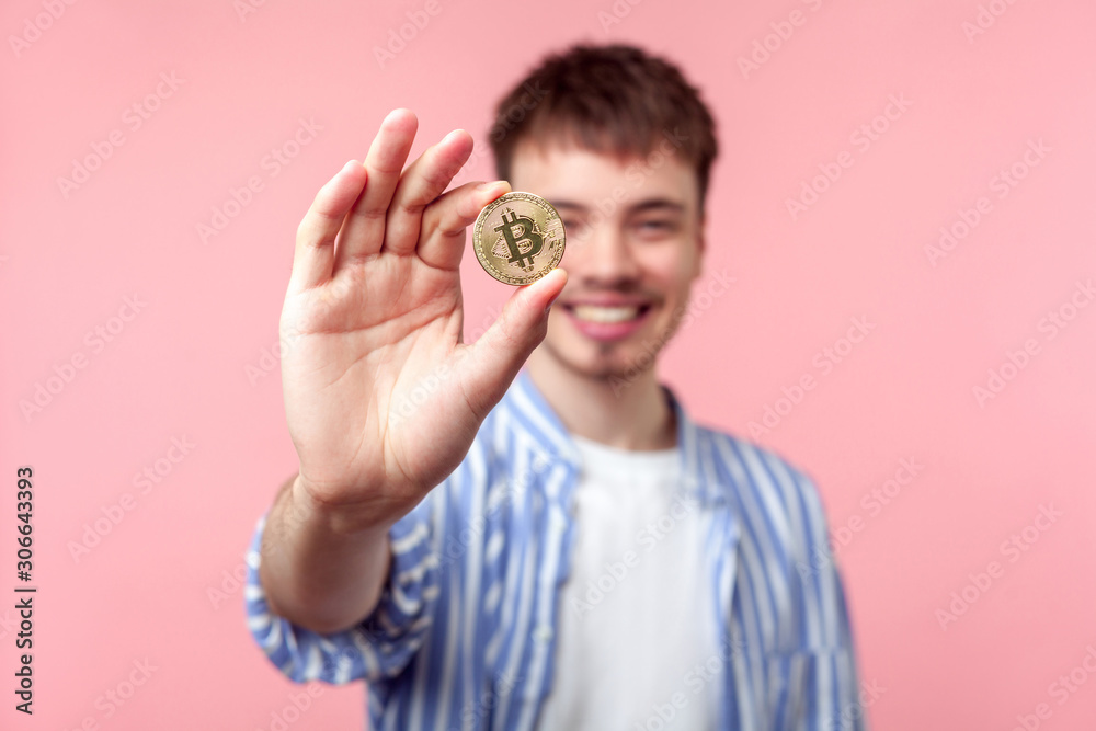 Fototapeta premium Portrait of positive excited brown-haired man in casual shirt standing showing golden bitcoin and looking at camera with toothy smile, cryptocurrency. indoor studio shot isolated on pink background