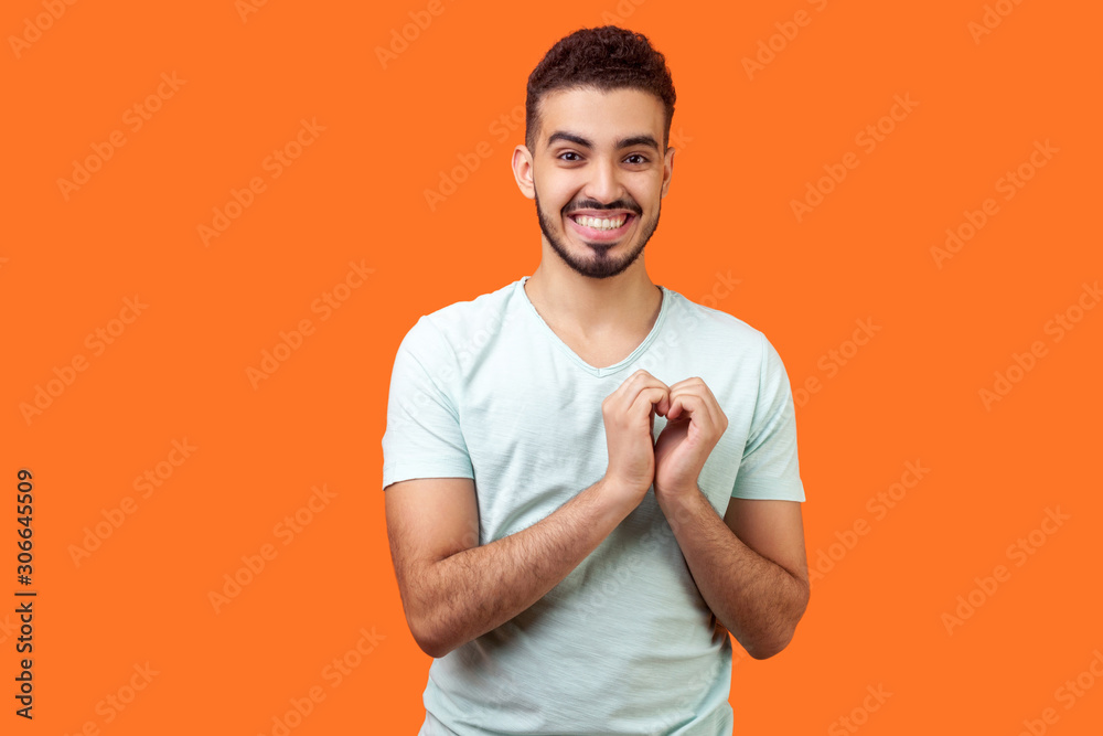 Portrait of joyous handsome brunette man in love making heart gesture with hands and smiling broadly at camera, demonstrating romantic feelings. indoor studio shot isolated on orange background Portrait of joyous handsome brunette man in love making heart gesture with hands and smiling broadly at camera, demonstrating romantic feelings. indoor studio shot isolated on orange background