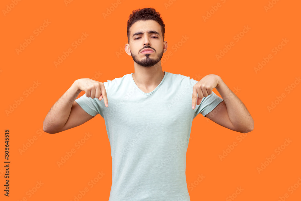 Here and right now. Portrait of bossy strict brunette man with beard in white t-shirt pointing down and looking arrogant, demanding to do immediately. indoor studio shot isolated on orange background Here and right now. Portrait of bossy strict brunette man with beard in white t-shirt pointing down and looking arrogant, demanding to do immediately. indoor studio shot isolated on orange background