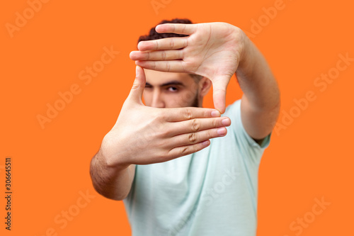 Portrait of attentive curious brunette man in casual white t-shirt looking at camera with one eye, focusing through photo frame made of fingers. indoor studio shot isolated on orange background photo