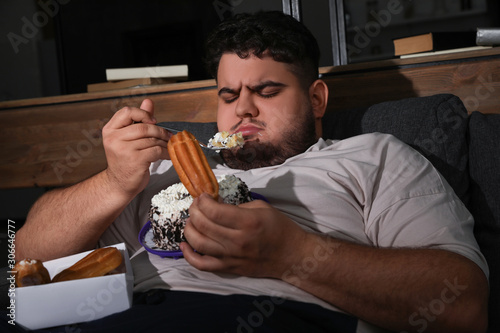 Depressed overweight man eating sweets in living room at night