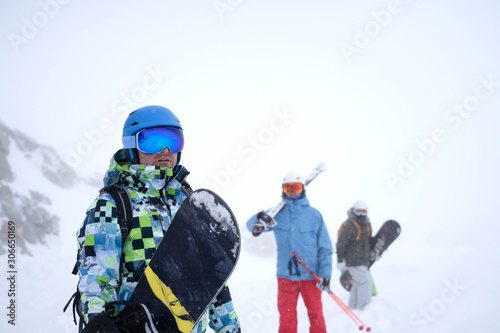 Photo of three sports people with skis and snowboard walking through winter resort