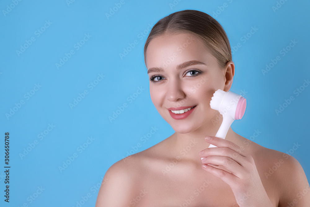 Young woman washing face with cleansing brush on light blue background. Cosmetic product Young woman washing face with cleansing brush on light blue background. Cosmetic product
