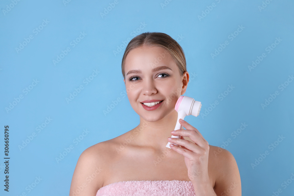 Young woman washing face with cleansing brush on light blue background. Cosmetic product