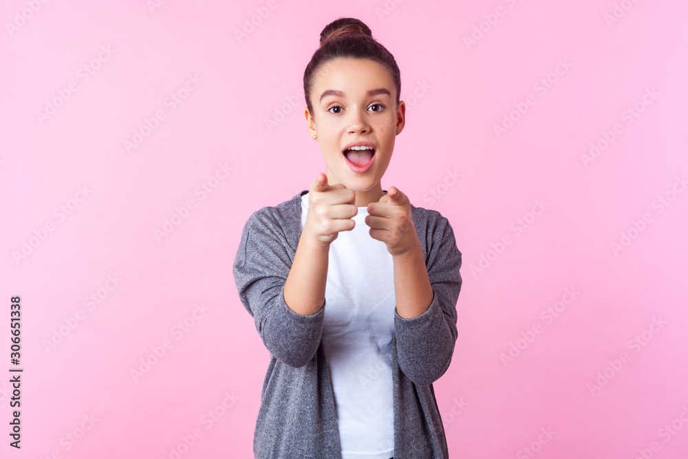 Hey you! Portrait of surprised brunette teenage girl with bun hairstyle in casual clothes pointing at camera, standing with open mouth, wondered astonished expression. studio shot, pink background Hey you! Portrait of surprised brunette teenage girl with bun hairstyle in casual clothes pointing at camera, standing with open mouth, wondered astonished expression. studio shot, pink background