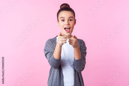 Hey you  Portrait of surprised brunette teenage girl with bun hairstyle in casual clothes pointing at camera  standing with open mouth  wondered astonished expression. studio shot  pink background