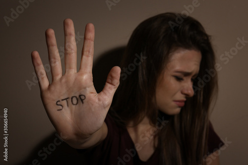 Abused young woman showing palm with word STOP near beige wall, focus on hand. Domestic violence concept