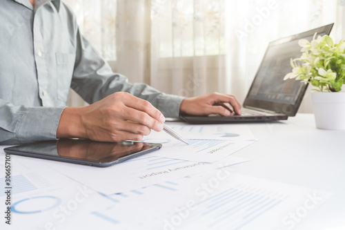 Asian businessman is using a pen to write a document report with laptop, tablet on a desk in the office. Staff checking the paper and planning the company's finances