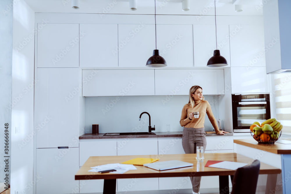 Attractive caucasian blonde fashionable woman leaning on kitchen counter, holding mug with coffee and looking trough window. Attractive caucasian blonde fashionable woman leaning on kitchen counter, holding mug with coffee and looking trough window.