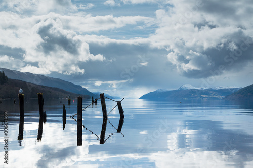 A view across Loch Ness with a broken pier in the foreground, and the length of the lake in the background and dark clouds above, in Scotland, UK
