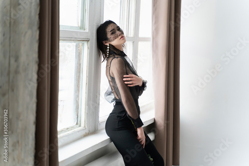 A beautiful Asian girl wearing trousers, a transparent blouse and a veil is standing in a bright room by the window. Advertising, commercial, fashion design.