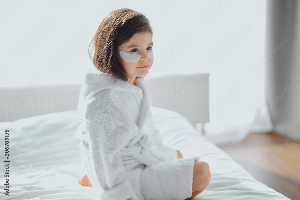 cute lovely girl in bathrobe sit on bed with facial mask for eyes, looking side in white room cute lovely girl in bathrobe sit on bed with facial mask for eyes, looking side in white room