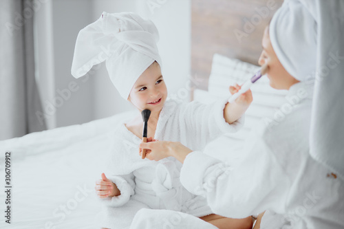 caucasian woman and little daughter sit on bed wearing towels and bathrobe doing makeup after shower. family concept