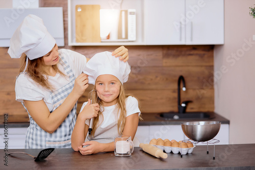 caucasian adult woman with dreamy chld in kitchen. mother prepare daughter to bake or cook while her child smile and think holding mixer , fixing cap, wearing apron
