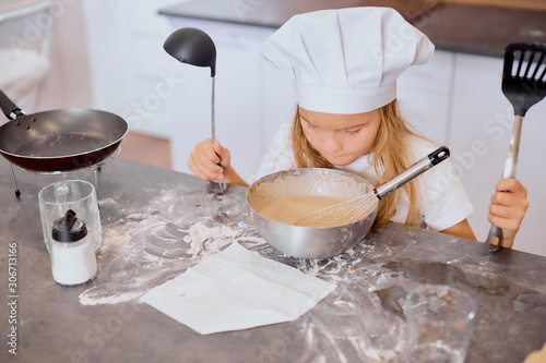 little baker girl interested in baking and cooking, look at dough in bowl, wearing apron and white cap for cooking, holding scapulas isolated in kitchen