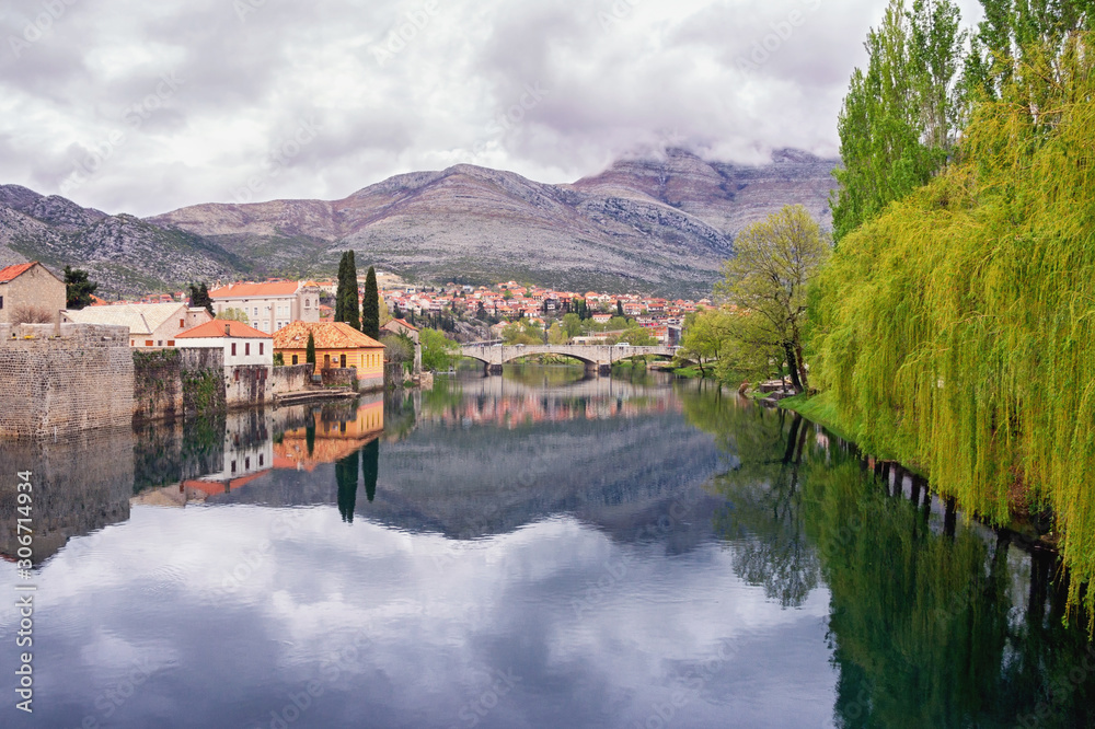 Cloudy spring day. Clouds, mountains, green trees and town are reflected in water of river. Bosnia and Herzegovina. View of Trebisnjica river and Old Town of Trebinje