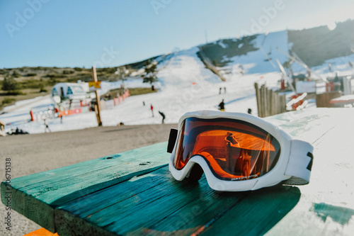 detail of some snowboard goggles on a green table in a ski resort full of snow and people