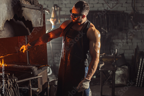 male blacksmith forging in his workshop, make iron products for manifacture of fireplaces and stoves. Use furnace for heating metal. Wearing protective eyeglasses and gloves