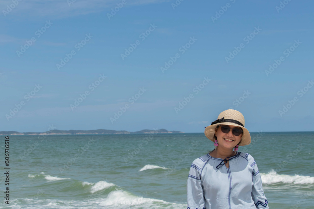 Portrait asian women wear a cream-colored knit hat, tied with black cloth, wear sunglasses, wear blue long-sleeved shirts, standing on a beach with a sea of water and an island far away.