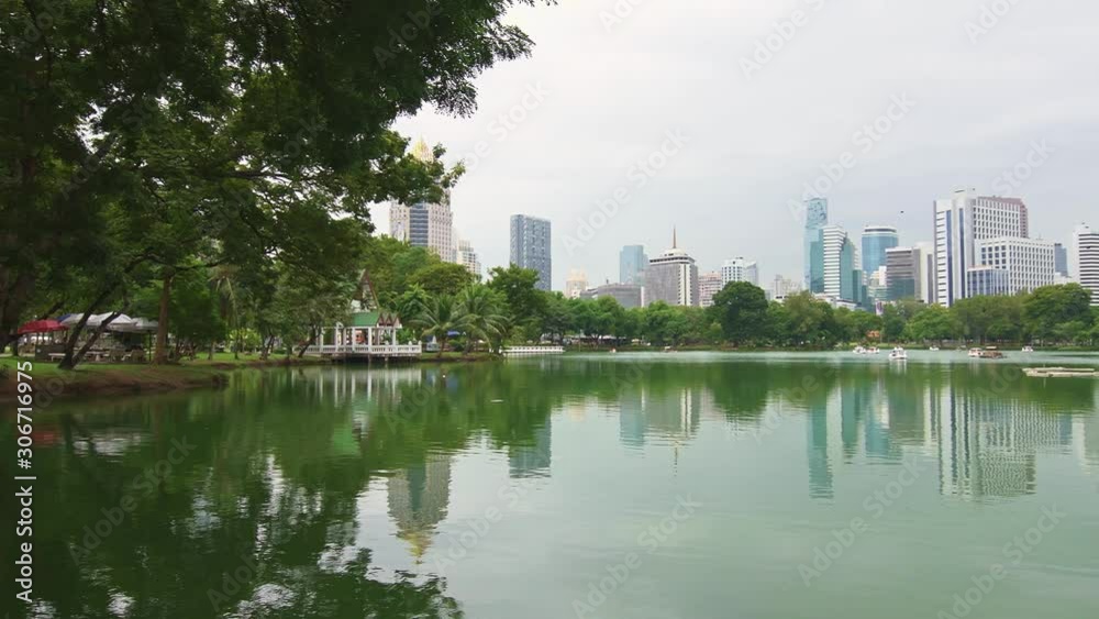 Central park Lumpini in Bangkok city, Thailand. Panoramic view of cityscape with skyscrapers and modern futuristic buildings. View of lake in the park with swan boat. Reflection in the water. Cloudy w Central park Lumpini in Bangkok city, Thailand. Panoramic view of cityscape with skyscrapers and modern futuristic buildings. View of lake in the park with swan boat. Reflection in the water. Cloudy w