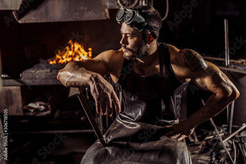 portrait of strong muscular bearded blacksmith or forger sit having rest after work, wearing leather apron and protective glasses for eyes on head, looking side