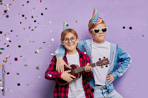 portrait of beautiful kid boys at happy birthday wearing holiday hat on head and holding ukulele, performing music. friendship, children, birthday concept, smile at camera