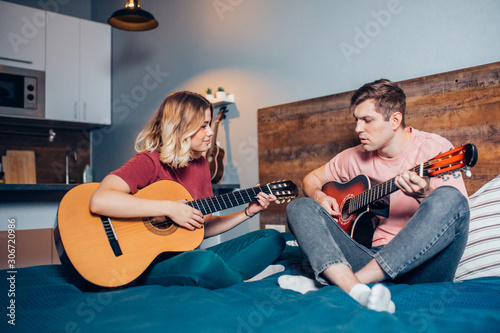 lovely couple, young people playing guitar together at home sitting on bed. caucasian girl and boy wearing casual home clothes
