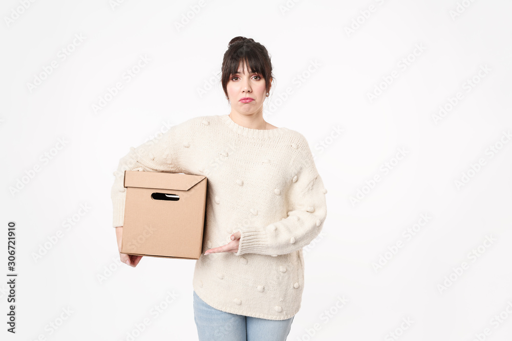 Sad and upset woman holding in armpit closed cardboard box with free copy space on grey background and pointing it by hand Moving day concept. ..packing unnecessary things at home. Sad and upset woman holding in armpit closed cardboard box with free copy space on grey background and pointing it by hand Moving day concept. ..packing unnecessary things at home.