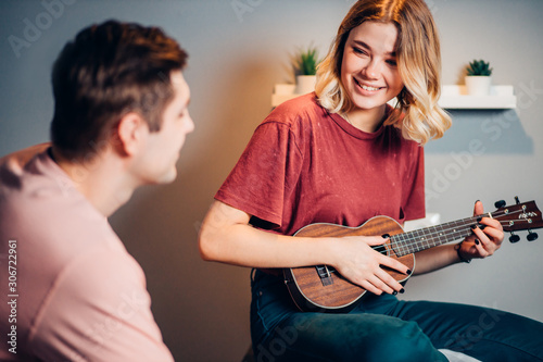 cute girl with blond short hair professionally play ukulele at home, young man look at process of performing music using ukulele