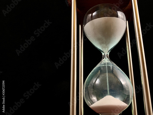 Hourglass on a stand, black background. Glass hourglass in the case. Glass time meter. Concept: time is running out, time management