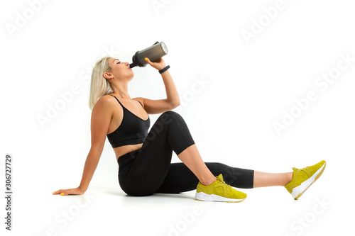 blonde girl in a black sports uniform drinks water sitting on the floor isolated white background