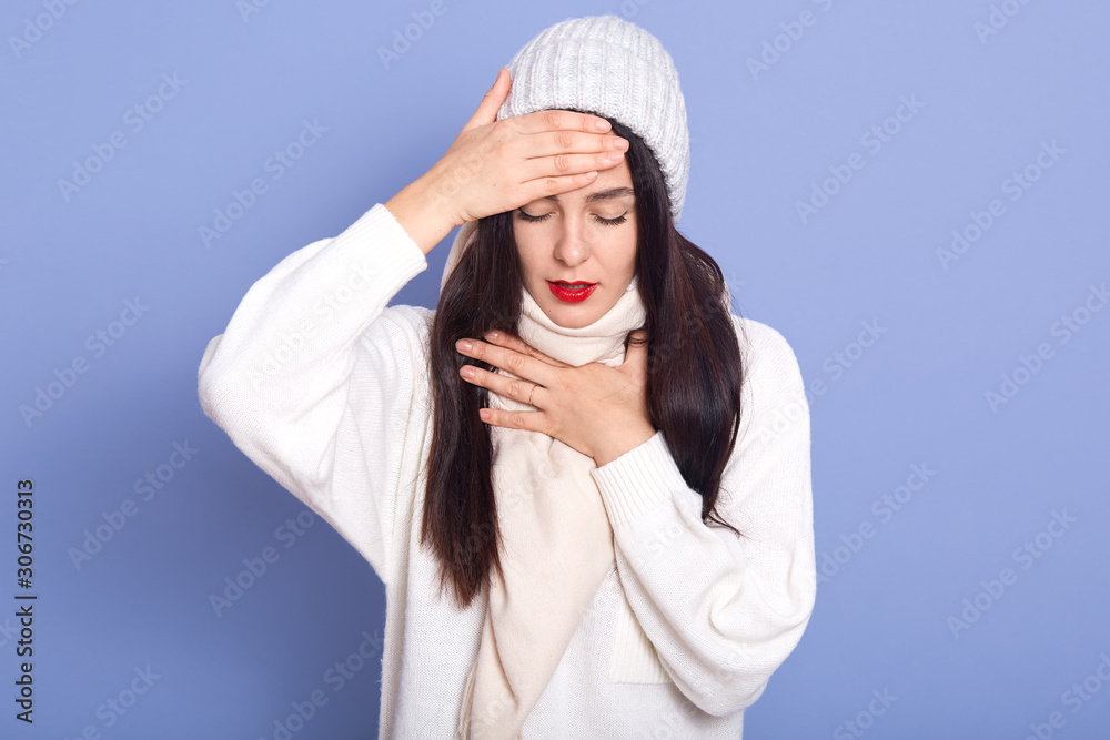 Image of upset attractive young girl closing her eyes, touching her forehead and neck, feeling unwell, having cold, running high temperature, wearing white hat, sweater and scarf. Health concept. Image of upset attractive young girl closing her eyes, touching her forehead and neck, feeling unwell, having cold, running high temperature, wearing white hat, sweater and scarf. Health concept.